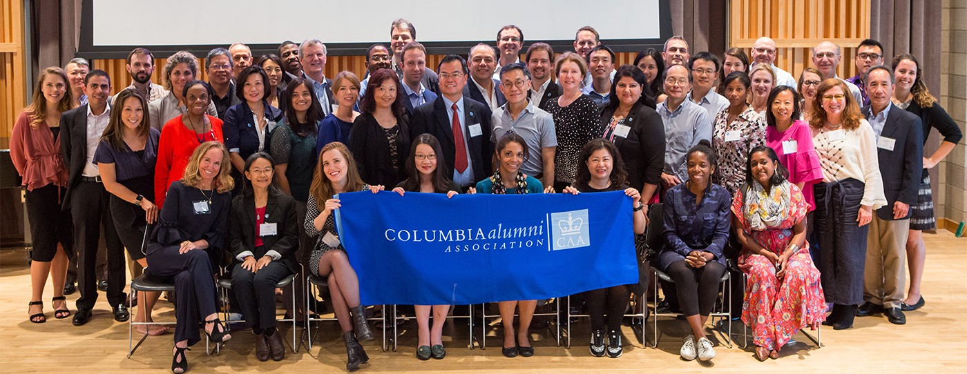 A group of around 50 men and women from all over the world smiling at the camera and holding a Columbia Alumni Association banner.